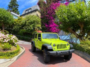 A bright green open-top sightseeing Jeep descending the lush, winding turns of Lombard Street in San Francisco, surrounded by green hydrangeas and brick paving.